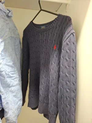 Mörkblå kabelstickad tröja Polo Ralph Lauren - Kabelstickad mörkblå tröja från Polo Ralph Lauren med rund halsringning och röd broderad logotyp på bröstet. Tröjan har långa ärmar och ribbade muddar vid ärmslut och nederkant. Perfekt för en stilren och klassisk look.
