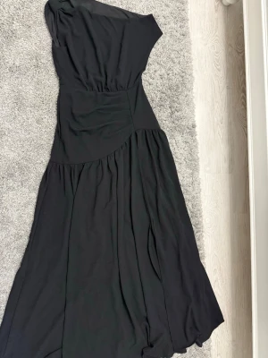 Svart oneshoulder långklänning - Snygg svart långklänning med oneshoulder-design och mjukt, fallande tyg. Klänningen har en asymmetrisk överdel och vida kjolpartier som ger en elegant siluett. Perfekt för dig som vill sticka ut med en stilren look.