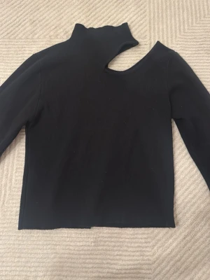 Svart oneshoulder topp med hög krage - Trendig svart topp med lång ärm och unik oneshoulder-design. Toppen har hög krage och en cool asymmetrisk öppning vid halsen. Perfekt för dig som vill sticka ut med en edgy look.
