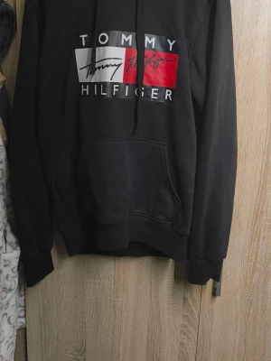 Svart hoodie från Tommy Hilfiger - Svart hoodie från Tommy Hilfiger med stor logga framtill i vitt, rött och svart. Klassisk känguruficka och justerbar huva med snören. Perfekt för en avslappnad och stilren look.