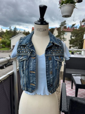 Kort jeansväst med nitar - Snygg kort jeansväst med coola silverfärgade nitar på axlar och övre rygg. Västen har två bröstfickor med lock och knappstängning framtill. Obs. Endast västen är till salu i denna annons. XXS-XS ganska liten. Gick ej att stänga på provdockan men man kan ju ha den öppen. 