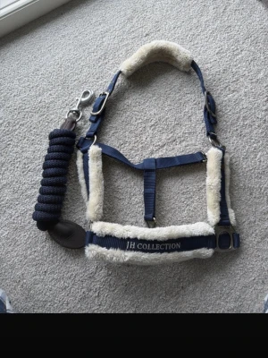 Blå grimma med ludd från JH Collection - Säljer en blå grimma med mjukt vitt ludd och matchande grimskaft från JH Collection. Grimman har justerbara spännen i metall och broderad logga. Perfekt för dig som vill ha snygga och bekväma hästtillbehör.