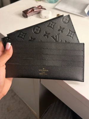  plånbok i läder - Snygg svart plånbok i äkta läder med klassiskt monogrammönster. Plånboken har flera kortfack och ett separat fack med dragkedja. Perfekt för att hålla koll på kort och kontanter med stil.