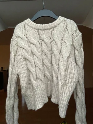 Kabelstickad tröja - Säljer en grå/beige kabelstickad tröja med breda flätmönster framtill och på ärmarna. Tröjan har rund halsringning och ribbade muddar vid ärmslut och nederkant. 