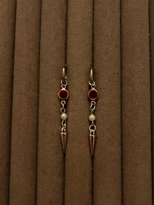 Silvriga örhängen med röd sten och pärla - Snygga örhängen i silverfärg med runda röda stenar, små vita pärlor och spetsiga hängen. Perfekt för dig som gillar unika detaljer och en edgy stil. Materialet ser ut att vara metall med dekorativa stenar och pärlor.