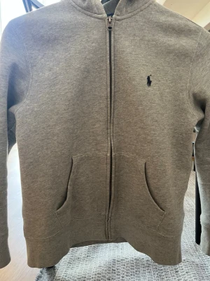Grå ziptröja från Ralph Lauren - Klassisk grå ziptröja från Ralph Lauren med den ikoniska broderade loggan på bröstet. Tröjan har dragkedja framtill och är tillverkad i mjuk bomull. Perfekt för lager på lager och enkel att matcha med olika outfits.