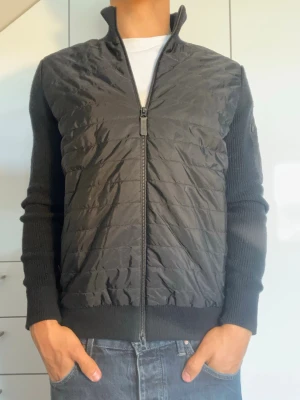 Svart cardiganjacka från J.Lindberg - Svart cardigan från J.Lindberg. Strl M, det finns både en innerficka och en ficka på vänsterarmen( se bild 5). Modellen är 178 cm och väger 77 kg. Hör gärna av dig vid frågor!