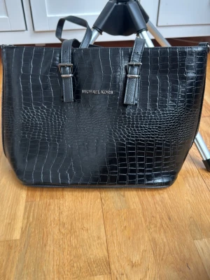 Svart croco-handväska Michael Kors - Snygg svart handväska från Michael Kors i croco-mönstrat konstläder. Väskan har två justerbara axelremmar med silverfärgade spännen och dragkedja upptill. Perfekt för dig som gillar stilrena och trendiga accessoarer.