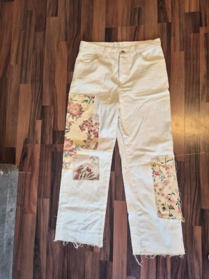 Vita jeans med blommiga lappar - använda 1 gång - Unika vita jeans med raka ben och råa benslut, dekorerade med stora blommiga lappar i olika färger. Jeansen har klassisk femficksdesign och hög midja. Perfekta för dig som vill sticka ut med en kreativ och personlig stil. Från Mango, använda enbart kort 1 gång. Storlek 38. 