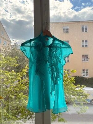 Turkos blus med volanger Saint Tropez - Snygg turkos blus från Saint Tropez i transparent, lätt material. Blusen har korta ärmar och dekorativa volanger längs knäppningen framtill. Perfekt för dig som vill sticka ut med färg och detaljer.