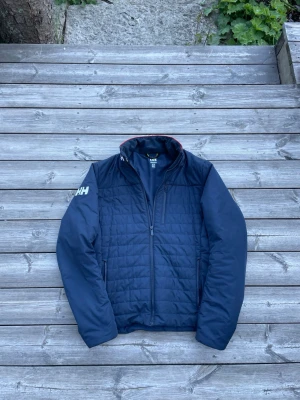 Mörkblå vindjacka från Helly Hansen - Snygg mörkblå vindjacka från Helly Hansen med quiltad front och släta ärmar. Jackan har dragkedja, hög krage och diskret HH-logga på ärmen. Perfekt för blåsiga dagar och enkel att matcha med olika outfits.