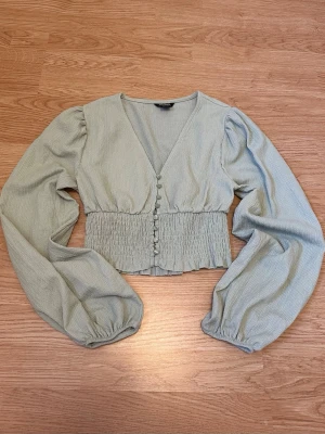 Ljusgrön topp - Ljusgrön topp med v-ringning, puffiga långärmar och smockad midja. Knäppning framtill. Monki. Strl S