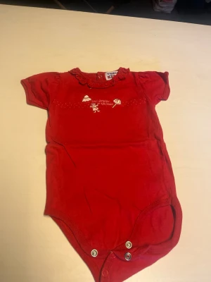 Röd body med volangkrage och tryck - Söt röd kortärmad body med volangkrage och små tryckknappar i grenen. Framtill finns ett gulligt broderat motiv och text. Perfekt för en mysig och lekfull stil.