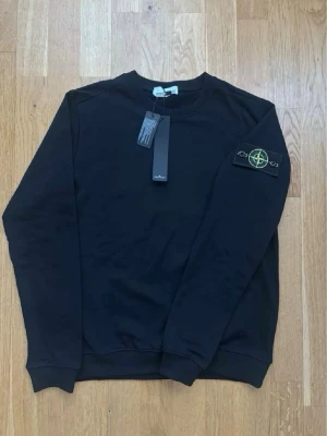 Svart sweatshirt från Stone Island - Svart sweatshirt från Stone Island med klassisk rund hals och ribbade muddar. Ikonisk logotyp-patch på vänster ärm. Tröjan är i mjukt material och har en stilren, enkel design som passar perfekt till streetwear.