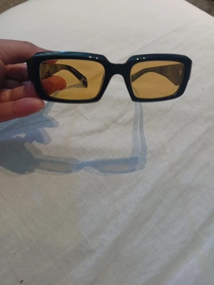 Svarta Prada solglasögon med gula glas - Snygga solglasögon från Prada med svart, kantig båge och gula glas. Modellen har en modern, rektangulär form och tydlig Prada-logga på sidan. Perfekta för dig som vill sticka ut med en cool accessoar.
