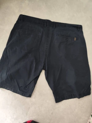 Mörkblå bomullsshorts från 157 - Säljer ett par mörkblå shorts från 157 med klassisk design. De har normal passform, sidofickor och bakfickor med knapp. Materialet är mjuk bomull och shortsen har bälteshällor samt knapp och dragkedja framtill. Perfekta för sommarens alla äventyr.