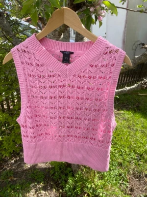 Rosa stickad väst från Lindex - Säljer en söt rosa stickad väst från Lindex med V-ringning och hålmönster. Västen är ärmlös och har bred ribbad kant nedtill och vid halsen. Perfekt att styla över en skjorta eller t-shirt för en trendig look.