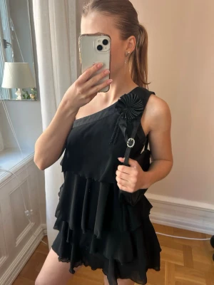 One shoulder mini-dress✨ - Svart kortklänning med one shoulder-design och flera lager volanger. Trendig och perfekt till festligheter✨Dekorativ blomdetalj på axeln. Ger snygg siluett. 