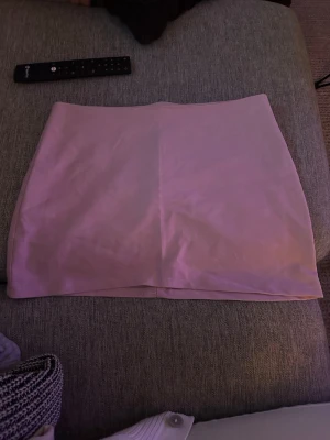 Ljusrosa kortkjol i syntetmaterial - Säljer en stilren ljusrosa kortkjol med rak passform. Kjolen har en diskret dragkedja bak och är tillverkad i ett slätt syntetmaterial. Perfekt att matcha med en enkel topp eller stickad tröja.