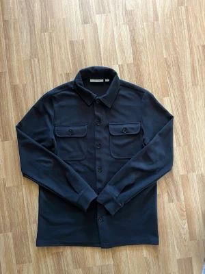 Svart overshirt från Only & sons - Stilren svart overshirt från Only & sons med klassisk krage och fyra knappar framtill. Två stora bröstfickor med lock och knapp. Tillverkad i ett strukturerat tyg som ger en modern känsla. Perfekt att bära som lager över t-shirt eller hoodie.