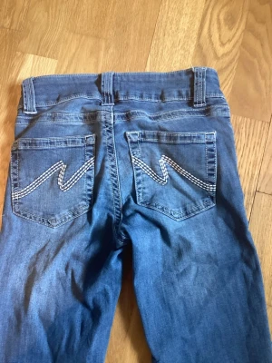 Blå bootcut jeans med broderade fickor - Snygga blå bootcut jeans med två knappar i midjan och broderade bakfickor med zigzag-mönster. Klassisk femficksmodell och normalhög midja. Jeansen är uppsydda till 75 cm innebenslängd. Strl 32