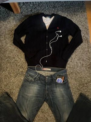 Polo Ralph Lauren Cardigan - Mörkblå kabelstickad kofta från Polo Ralph Lauren med V-ringning och knappar framtill. Klassisk röd logga broderad på bröstet. Långa ärmar och ribbade muddar. Perfekt för lager-på-lager och snygg till jeans.
