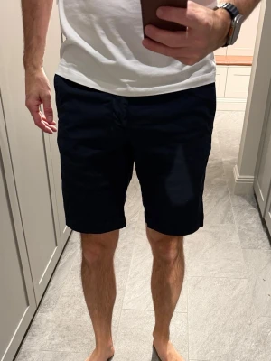 Mörkblå shorts  - Klassiska mörkblå shorts i bomull med slimfit passform. De har fickor på sidorna, bälteshällor och stängs med knapp och dragkedja. Perfekta för sommaren och enkla att matcha med olika outfits.