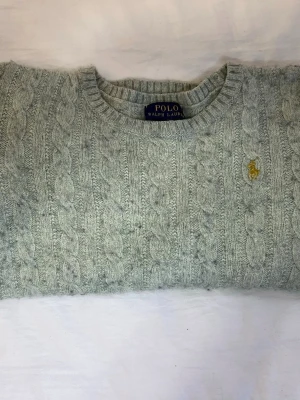 Ljusgrå kabelstickad tröja Polo Ralph Lauren - Kabelstickad tröja i ljusgrått från Polo Ralph Lauren med klassisk rund halsringning och broderad gul logotyp på bröstet. Tröjan är gjord i ett mjukt och värmande material, perfekt för kyliga dagar. Snygg och tidlös design som passar till det mesta.