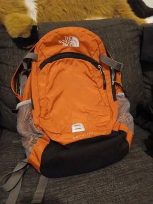 Orange ryggsäck från The North Face - Snygg orange ryggsäck från The North Face med svarta och grå detaljer. Ryggsäcken har flera fack med dragkedja, meshfickor på sidorna och vadderade axelremmar för extra komfort. Perfekt för skola eller äventyr. Materialet är slitstark nylon.