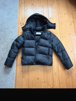 Svart pufferjacka från Calvin Klein - Snygg svart pufferjacka från Calvin Klein med avtagbar huva och diskret logga på bröstet. Jackan är vadderad och har en boxig passform med dragkedja framtill. Perfekt för kalla dagar och riktigt varm tack vare sitt tjocka material.