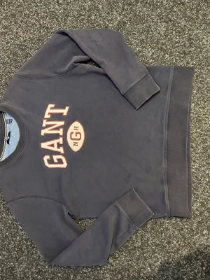 Mörkblå sweatshirt från GANT - Mörkblå sweatshirt från GANT med klassisk logga i vitt och rött framtill. Tröjan har rund hals, ribbade muddar vid ärmslut och nederkant samt mjukt material på insidan. Perfekt för en avslappnad och sportig stil. Kolla gärna in mina andra annonser