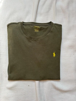 Olivgrön t-shirt från Polo Ralph Lauren - Olivgrön t-shirt från Polo Ralph Lauren med klassisk gul broderad logga på bröstet. T-shirten har rund halsringning och korta ärmar. Tillverkad i mjuk bomull för en skön känsla och stilren look. Inga hål eller andra defekter. Strolek M passar även S. Bara höra av sig vid minsta fundering😊