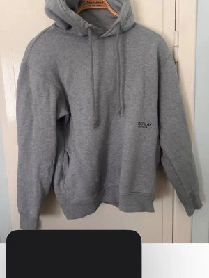 Grå hoodie från Replay - Klassisk grå hoodie från Replay med huva och dragsnören. Tröjan har en enkel design med liten Replay-logga på bröstet och en stor magficka. Tillverkad i mjukt bomullsmaterial och har ribbade muddar vid ärmslut och nederkant.