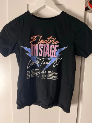 Svart t-shirt med Electric On Stage-tryck - Cool svart t-shirt med färgglatt tryck på bröstet där det står 'Electric On Stage World Tour 87' och en lista med turnédatum. T-shirten har rund hals och korta ärmar. Perfekt för dig som gillar retro och bandtröjor.