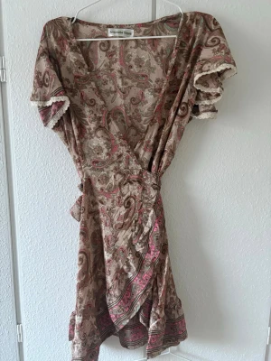 Beige omlottklänning med paisleymönster - Snygg kort omlottklänning från After Eden Store i beige med paisleyinspirerat mönster i rosa och brunt. Klänningen har volangdetaljer, knyt i midjan och korta ärmar. Perfekt för sommarens varma dagar.