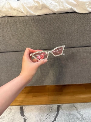 Rosa solglasögon med strassdetaljer - Snygga solglasögon med ljusrosa glas och kantiga bågar dekorerade med glittrande strass. Perfekt accessoar för att sticka ut och ge din outfit extra edge. Bågarna är i syntetmaterial och har en trendig, modern form.