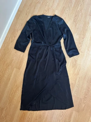 Svart omlottklänning - Svart klänning med omlott-design. Klänningen har trekvartsärm och är tillverkad i ett glansigt material för en elegant känsla