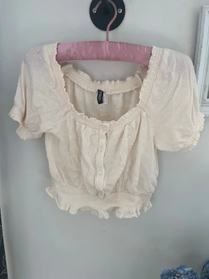 Krämvit kortärmad blus Gina Tricot - Blus i krämvit nyans från Gina Tricot med korta puffärmar, knappar framtill och volangkant vid nederdelen. Materialet är lätt och har ett diskret prickigt strukturmönster samt smockad midja som ger en feminin och trendig look.