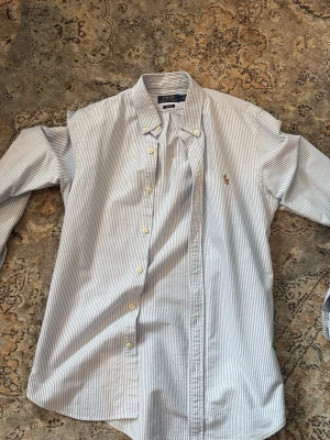 Randig skjorta från Polo Ralph Lauren - Klassisk blåvit randig skjorta från Polo Ralph Lauren i slim fit-modell. Skjortan har knappar framtill, button down-krage och broderad logga på bröstet. Tillverkad i bomull och har långa ärmar.
