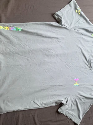 Grå Under Armour t-shirt - Snygg grå t-shirt från Under Armour med regnbågsfärgad logga och textdetaljer. Perfekt för träning eller en sportig look. Tillverkad i ett lätt och snabbtorkande material som andas.