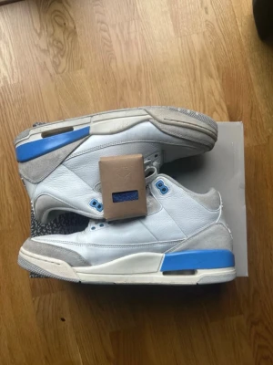 Jordan 3 lucky charm - Säljer ett par Air Jordan 3 Retro sneakers i vitt och blått med gråa detaljer. Skorna har klassisk snörning, ikonisk Jumpman-logga på hälen och är tillverkade i skinn och mocka. Perfekta för dig som gillar streetwear och vill sticka ut med en tidlös modell.