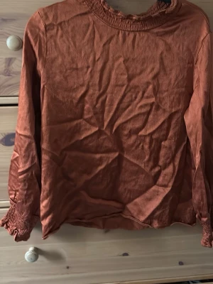 Rostfärgad blus med smockdetaljer - Trendig rostfärgad blus med lång ärm och smockdetaljer vid halsringning och ärmslut. Tillverkad i ett mjukt, lätt tyg som ger en avslappnad look. Perfekt att matcha med jeans eller kjol för en snygg outfit.