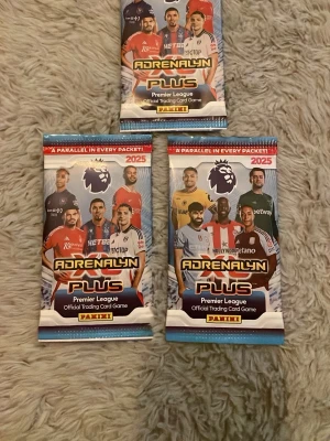 Adrenalyn Plus Premier League 2025 - Tre oöppnade booster packs Adrenalyn Plus Premier League 2025 från Panini. Varje paket innehåller officiella samlarkort med fotbollstema. Förpackningarna är färgglada i blått, rött och vitt med Premier League-loggan och bilder på spelare.