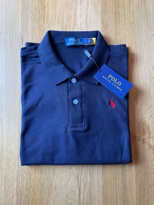 Marinblå pikétröja från Polo Ralph Lauren - Snygg marinblå pikétröja från Polo Ralph Lauren med klassisk krage, knappar framtill och ikonisk röd logga broderad på bröstet. Perfekt för en stilren och avslappnad look.
