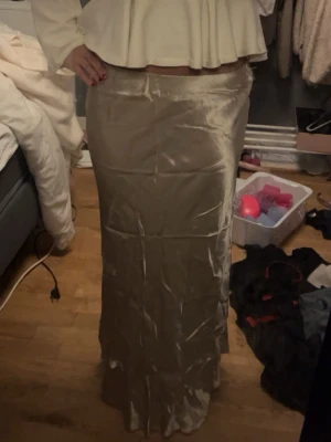 Champagnefärgad långkjol i satin - Snygg långkjol i champagnefärgat satinmaterial med glansig finish. Helt oanvänd men har blivit skrynklig av garderoben, men går att stryka så är den som ny
