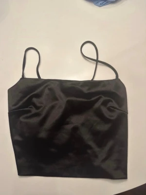 Svart satinlinnetopp med smala band - Snygg svart linnetopp i satin med smala axelband och snörning i ryggen. Toppen har en croppad passform och en stilren, minimalistisk look som passar perfekt till jeans eller kjol. Finns ingen lapp så vet ej storleken men gissar på S/M❤️