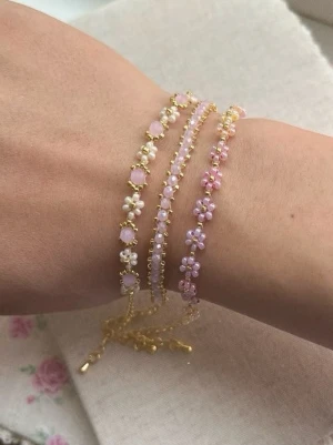 Söta armband med pärlor och blommor - Tre tunna armband i guldton med små pärlor och blommor i rosa och vitt. Armbanden har delikata kedjor och justerbar längd. Perfekt för dig som gillar romantiska och feminina accessoarer med en touch av pastell. 70kr/st
