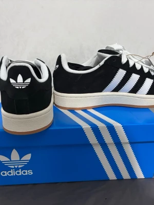 Adidas Campus svarta sneakers - Svarta Adidas Campus sneakers med klassiska vita ränder och vit sula. Ovandelen är i mocka och skorna har vita skosnören. Adidas logga syns på hälen och tungan. Snygg och tidlös modell som passar till många stilar.