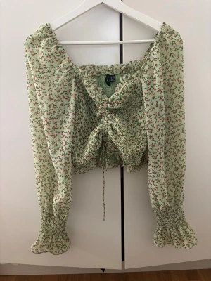 Grön blommig offshoulder-blus  - Söt ljusgrön offshoulder-blus med små röda blommor. Blusen har långa puffiga ärmar med smock vid ärmslut och midja, samt en rynkad detalj framtill med knytband. Perfekt för en trendig och romantisk look.