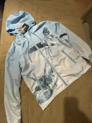 Trapstar vindjacka- windbreaker  - Snygg ljusblå vindjacka med huva och dragkedja framtill. Jackan har ett stort tryck av örnar i flykt på både fram- och baksidan samt textdetalj på ryggen. Två fickor med dragkedja och justerbar nederkant. Perfekt för dig som vill sticka ut.